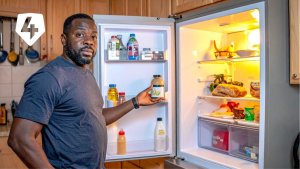 Man standing in front of an open refrigerator holding a jar of food with various items inside.